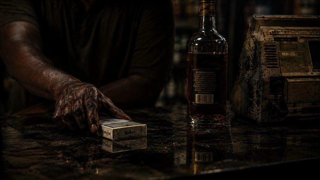 Close-up of a weathered hand reaching for a pack of cigarettes beside a whiskey bottle.